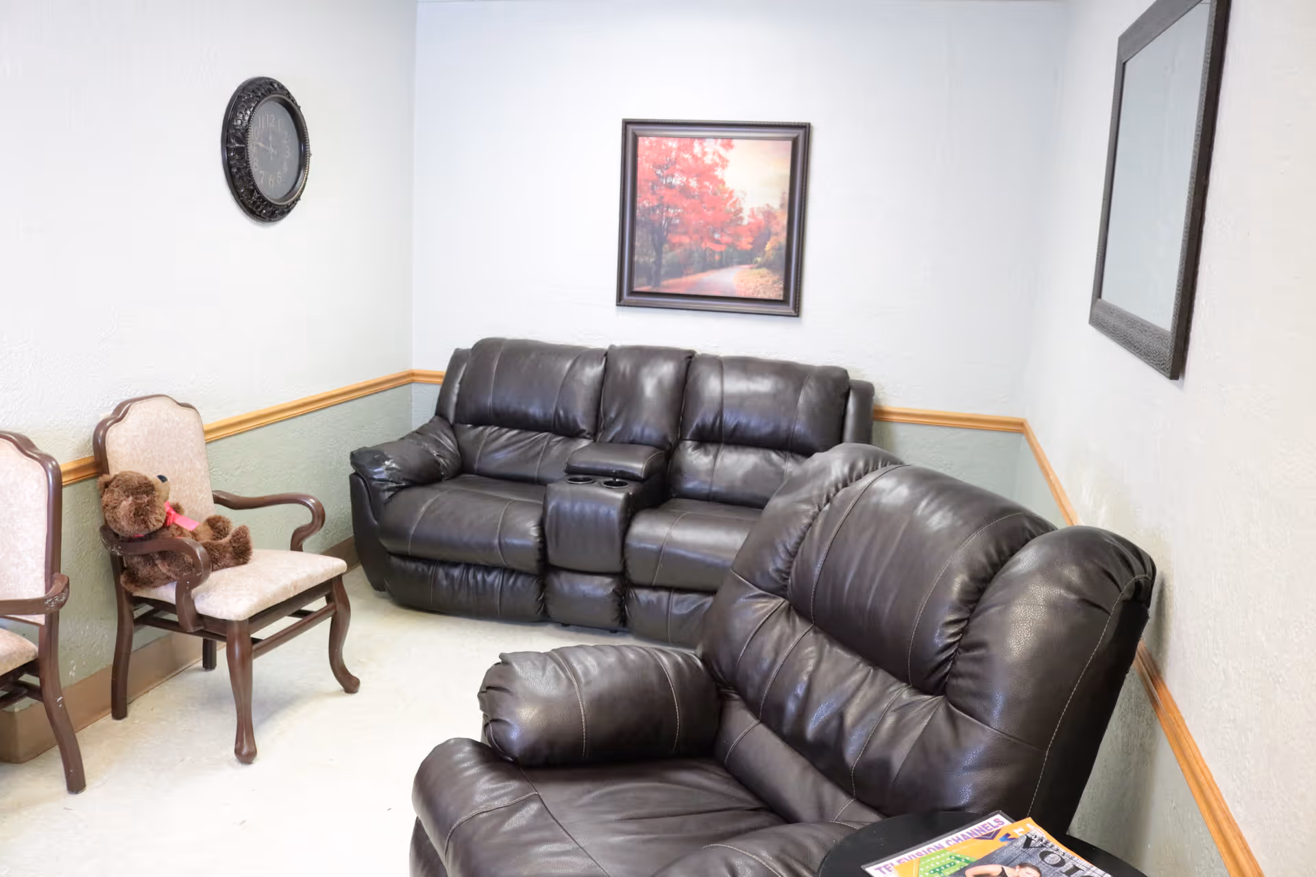 A cozy living room area with a black leather recliner and a matching black leather sofa. There are two wooden chairs with beige cushions, one of which has a brown teddy bear with a pink ribbon sitting on it. A framed picture of a tree-lined path with autumn foliage hangs on the wall above the sofa. A round wall clock is mounted on the adjacent wall. A small table next to the recliner holds a magazine.