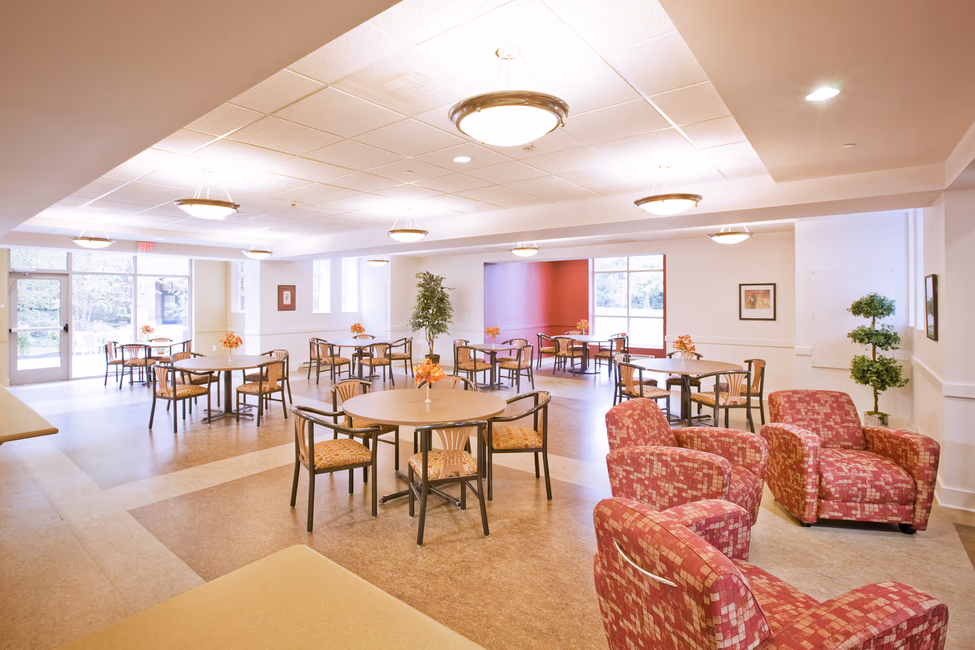 A spacious, well-lit dining room in Woodcrest Retirement Residence featuring multiple round tables with chairs arranged around them. Each table has a small floral centerpiece. There are also three red patterned armchairs grouped together near the foreground. Large windows and glass doors allow natural light to fill the room, and ceiling lights provide additional illumination. The walls are light-colored with some framed artwork and potted plants adding to the decor.
