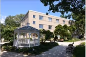 Outdoor view of a senior living facility with a white gazebo, benches, trees, and a multi-story beige building in the background under a clear blue sky.