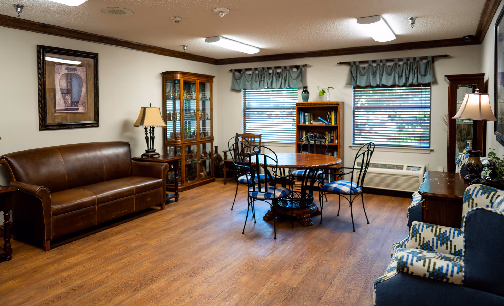 A cozy living room with wooden flooring, a brown leather sofa, a round wooden table with four metal chairs with blue cushions, two windows with blinds and blue valances, a wooden bookshelf, a glass display cabinet, two table lamps, and two patterned armchairs.