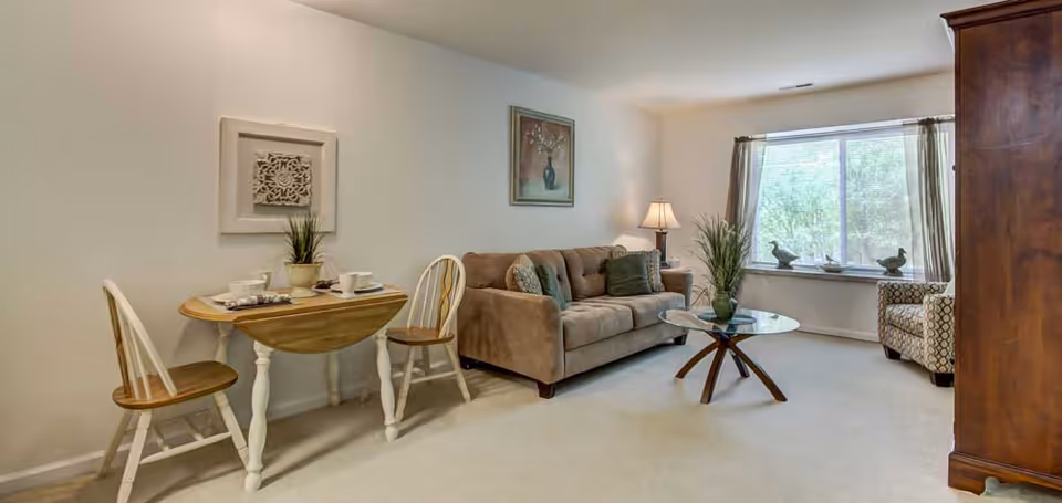 A cozy living room area featuring a brown sofa with decorative pillows, a glass coffee table with a plant centerpiece, a patterned armchair near a large window with curtains, and a small wooden dining table set with two chairs and tableware. The walls are decorated with framed artwork and a table lamp provides warm lighting.
