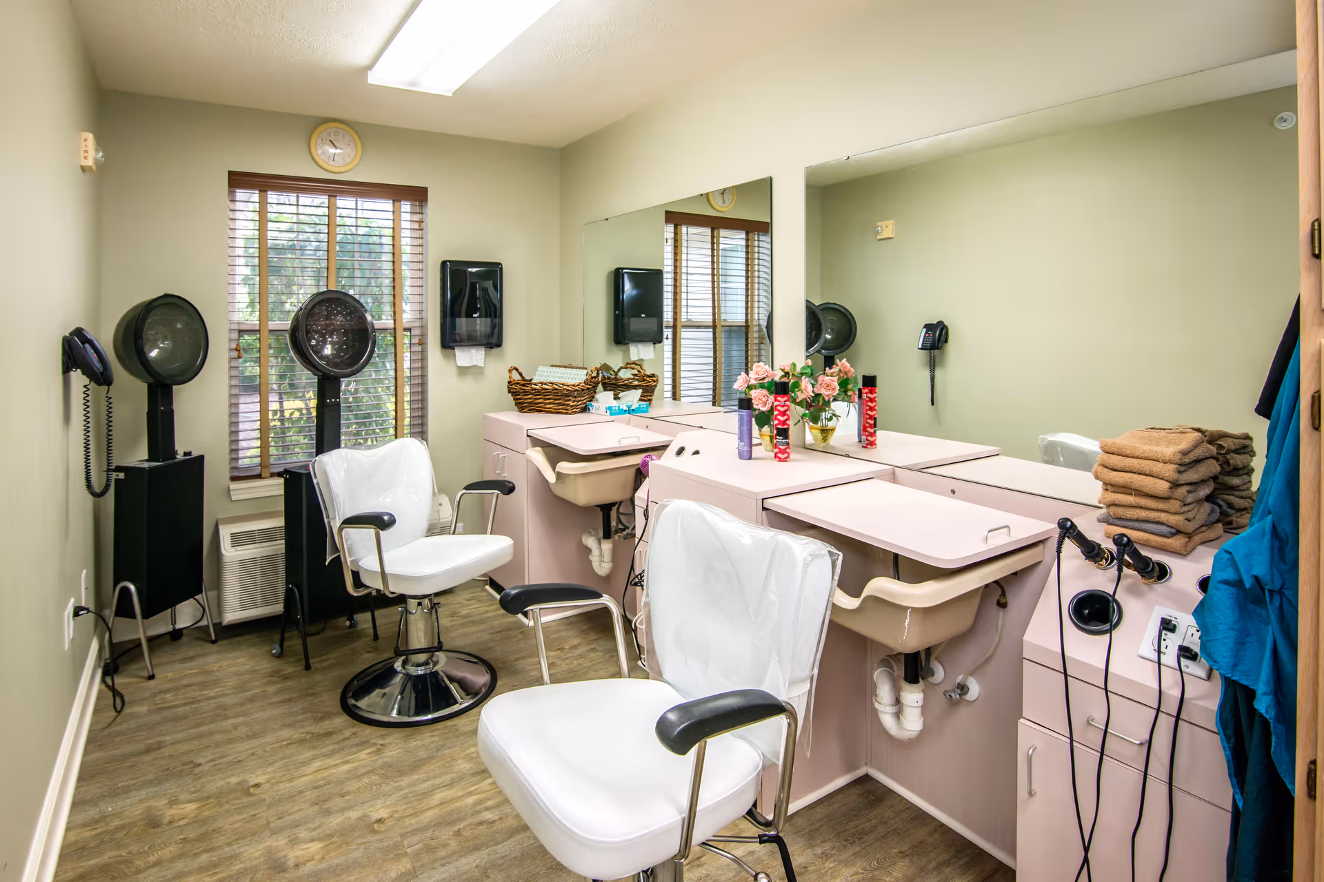 Interior view of a small salon area with two white salon chairs, hair dryers, a large mirror, a window with wooden blinds, and a counter with a sink, towels, and decorative flowers.