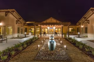 Night view of an outdoor courtyard at a senior living facility with a central decorative water fountain surrounded by rocks and plants, illuminated by pathway lights. The building structures with warm lighting are visible in the background under a dark sky.
