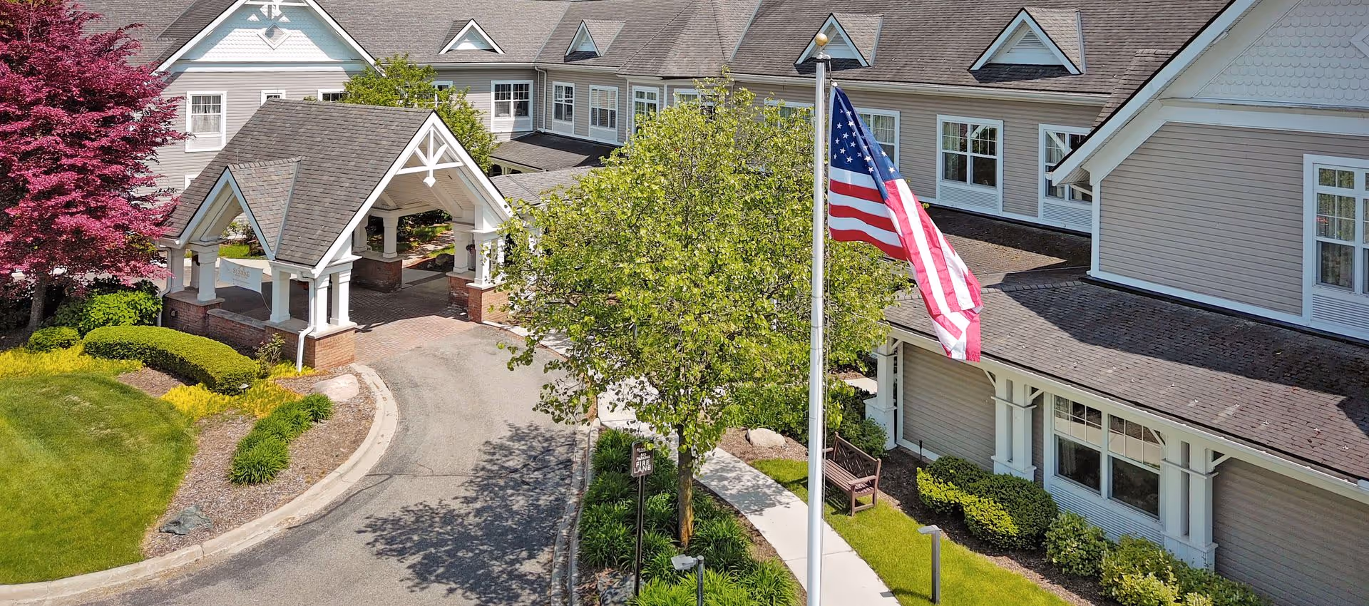 Front entrance of a senior living building with a covered porte-cochere, an American flag on a pole, and landscaped grounds.