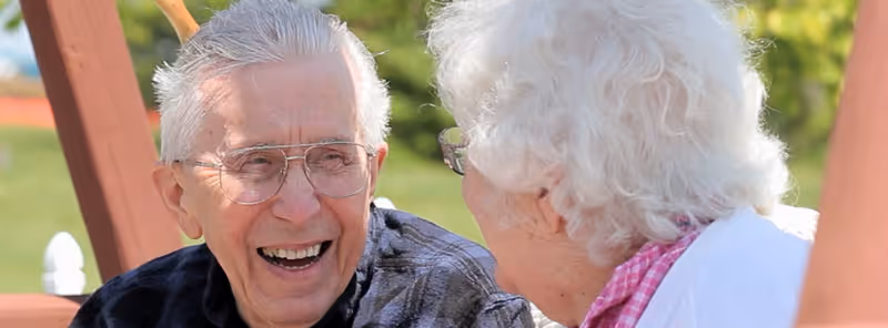 An elderly man and woman sitting outdoors on a wooden bench or swing, engaged in a joyful conversation. The man is smiling broadly and wearing glasses and a dark shirt, while the woman has white hair and is wearing glasses and a white top with a pink scarf. The background is blurred greenery.
