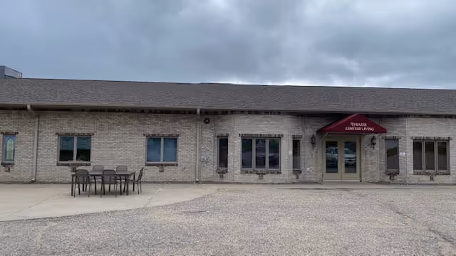 Exterior view of Frazee Assisted Living Home Care building with a brick facade, several windows, a maroon awning above the entrance door, and an outdoor table with chairs on the left side. The sky is cloudy.