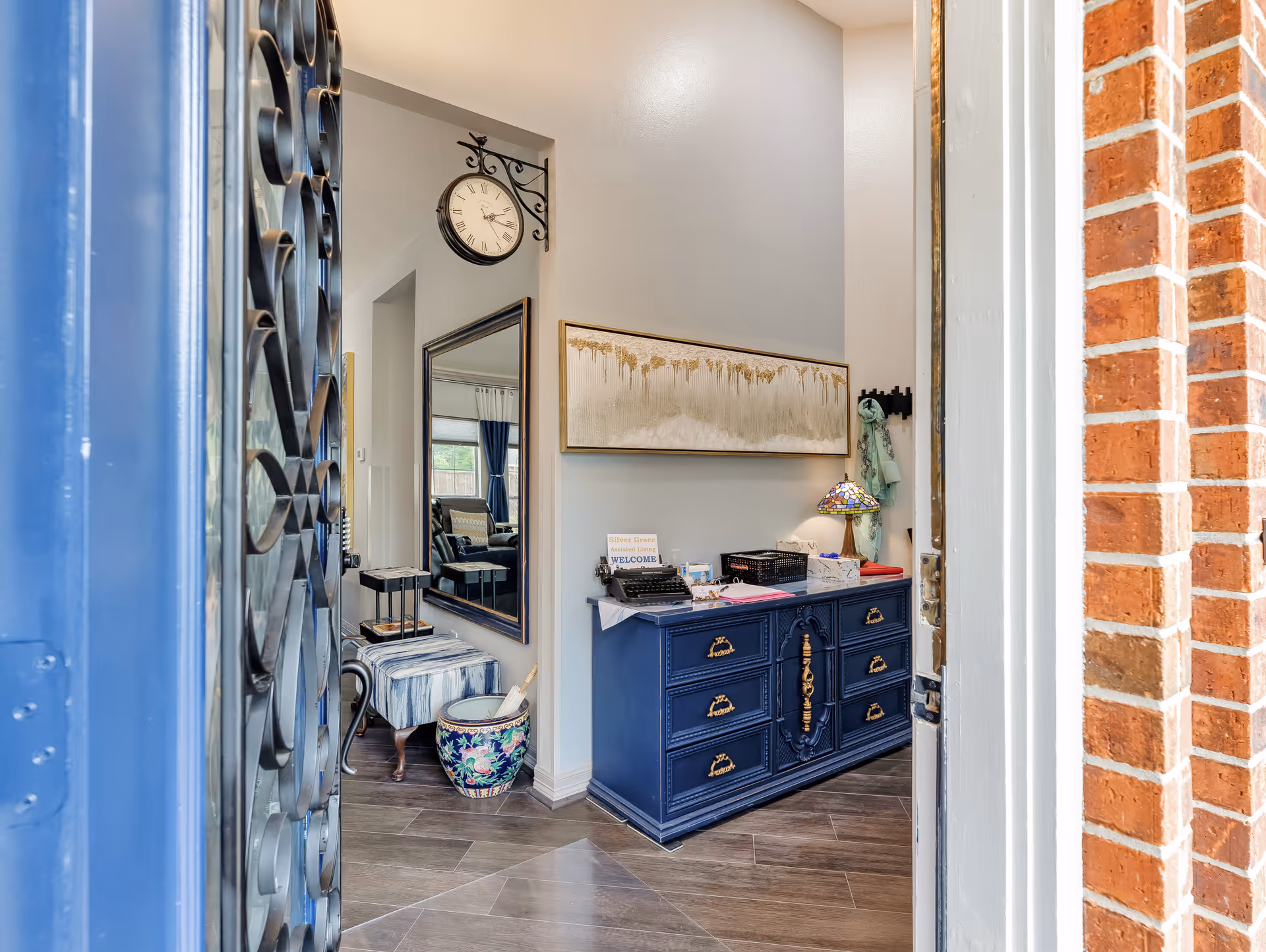 View through an open front door into a foyer with a blue dresser topped with a lamp and decor, a wall clock and mirror, and a brick doorway.