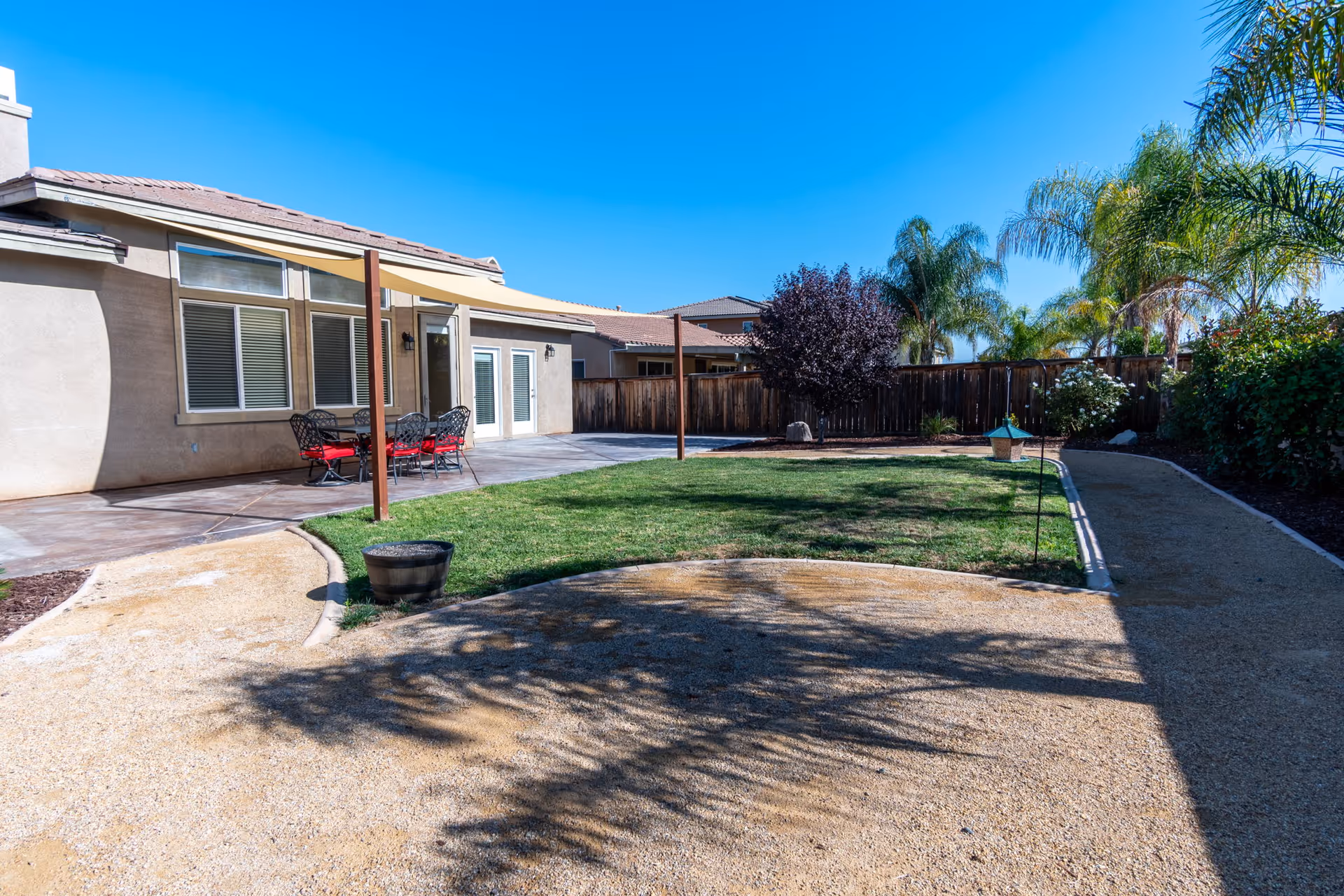 Outdoor patio area at Concord Estates Assisted Living featuring a concrete patio with a table and chairs with red cushions, a grassy lawn, a dirt walking path, palm trees, and a wooden fence under a clear blue sky.