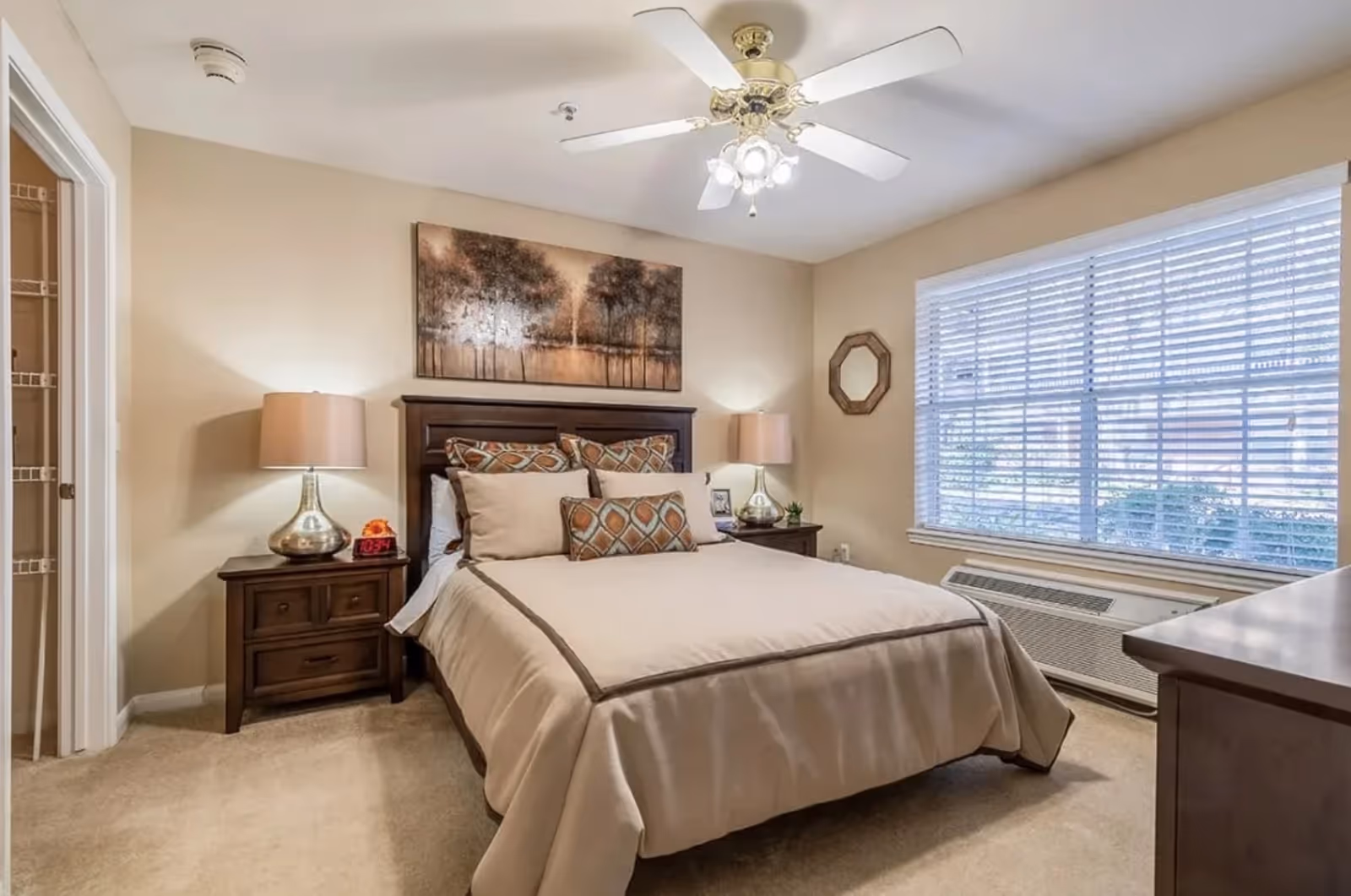 A cozy bedroom with a neatly made bed featuring beige and patterned pillows and a beige comforter. There are two wooden nightstands on either side of the bed, each with a lamp. A large window with white blinds lets in natural light. A ceiling fan with lights is mounted on the ceiling. A framed artwork of trees hangs above the bed, and a small decorative mirror is on the wall next to the window.