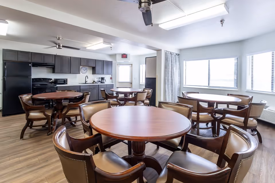 A bright communal dining area with several round wooden tables surrounded by leather-upholstered chairs. The room features a kitchen area with dark cabinets, a black refrigerator, stove, microwave, and coffee maker. Large windows with blinds allow natural light to fill the space, and ceiling fans and fluorescent lights are mounted on the white ceiling.