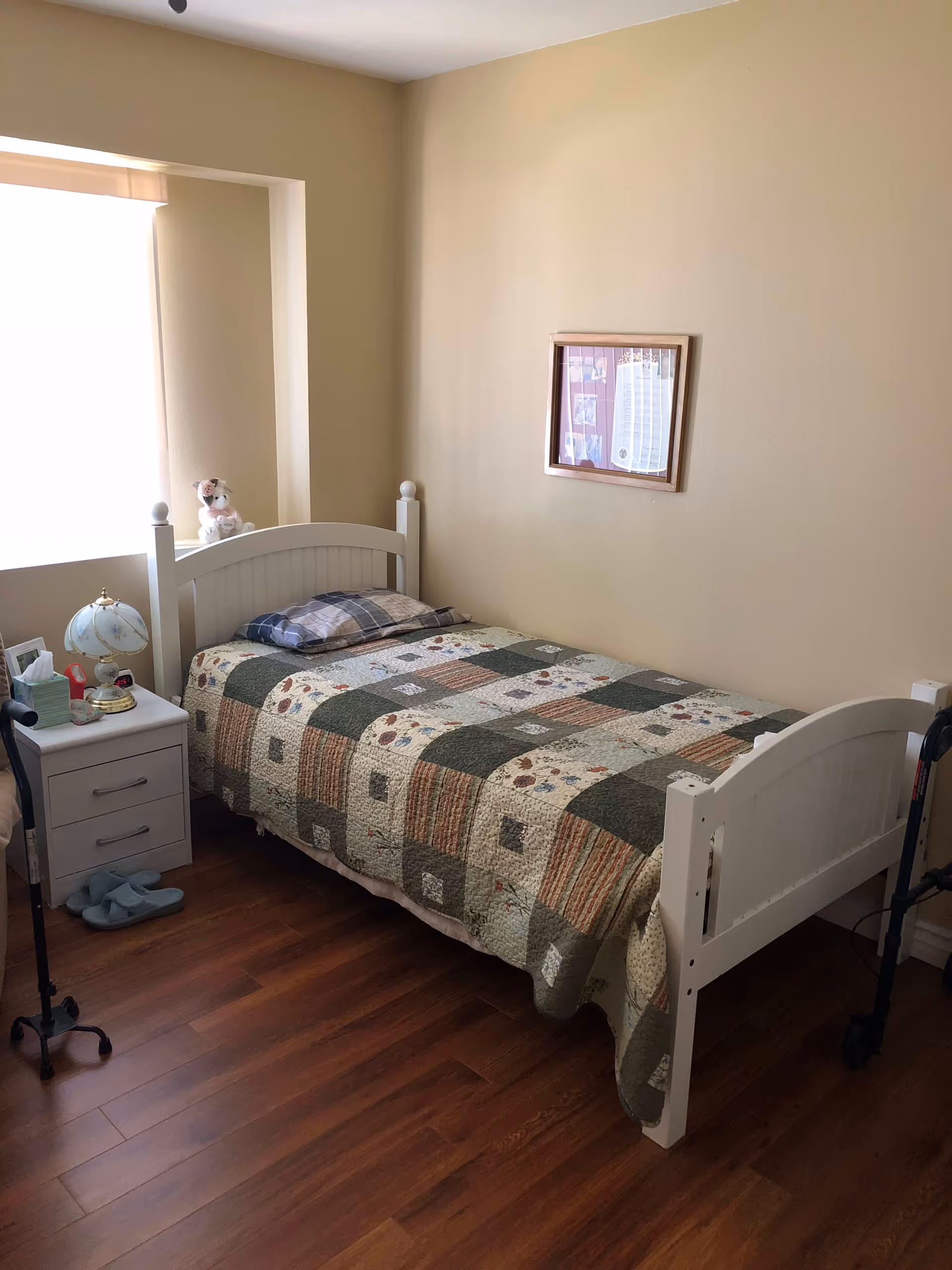 Small single bedroom with a white wooden bed covered by a patchwork quilt, a nightstand with a lamp, slippers on the floor and a mobility walker.