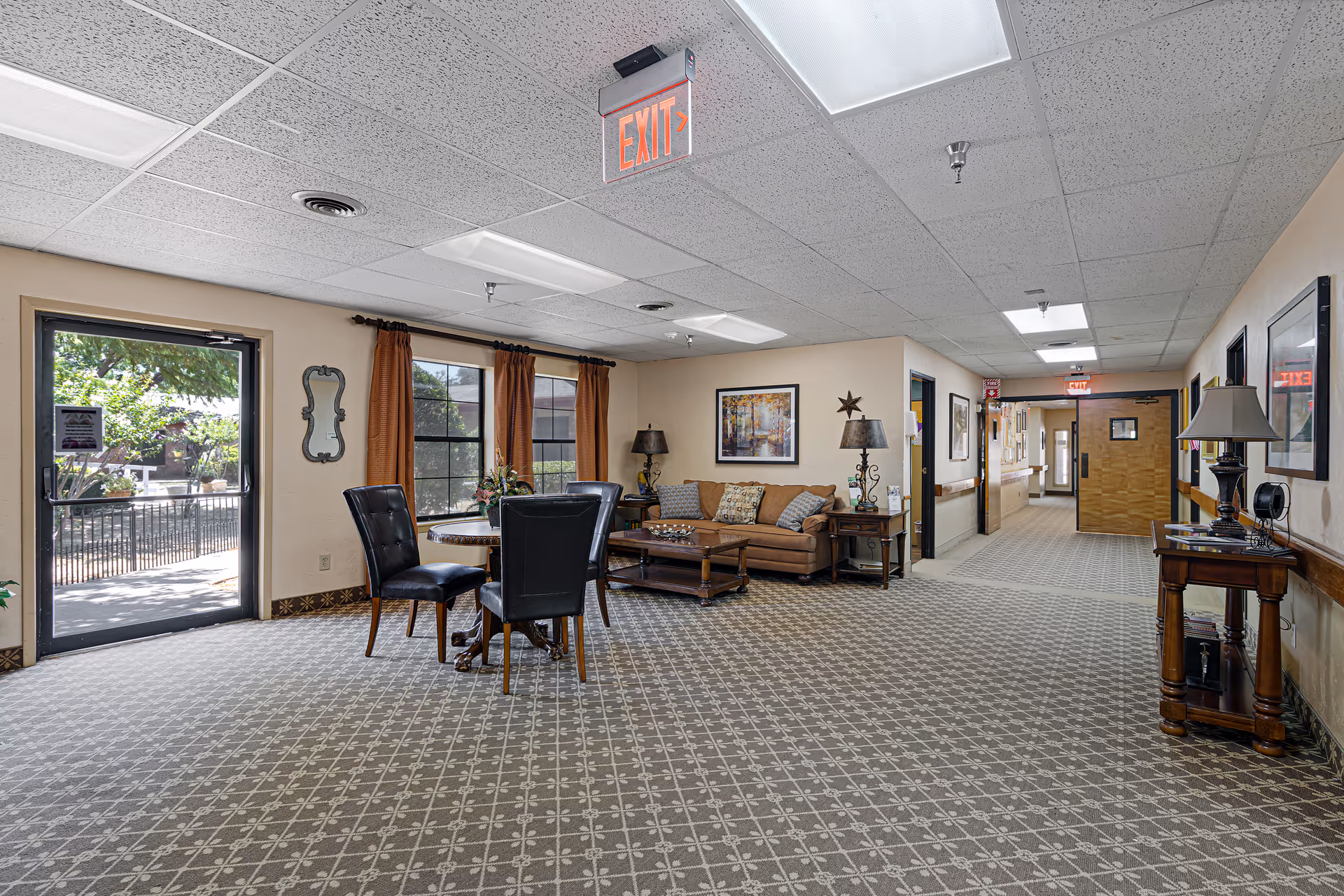 Interior view of a senior living facility lounge area with a patterned carpet, a small round table with four black chairs, a brown sofa with cushions, side tables with lamps, framed artwork on the walls, and large windows with brown curtains. There is an exit door leading outside and a hallway extending to the right.