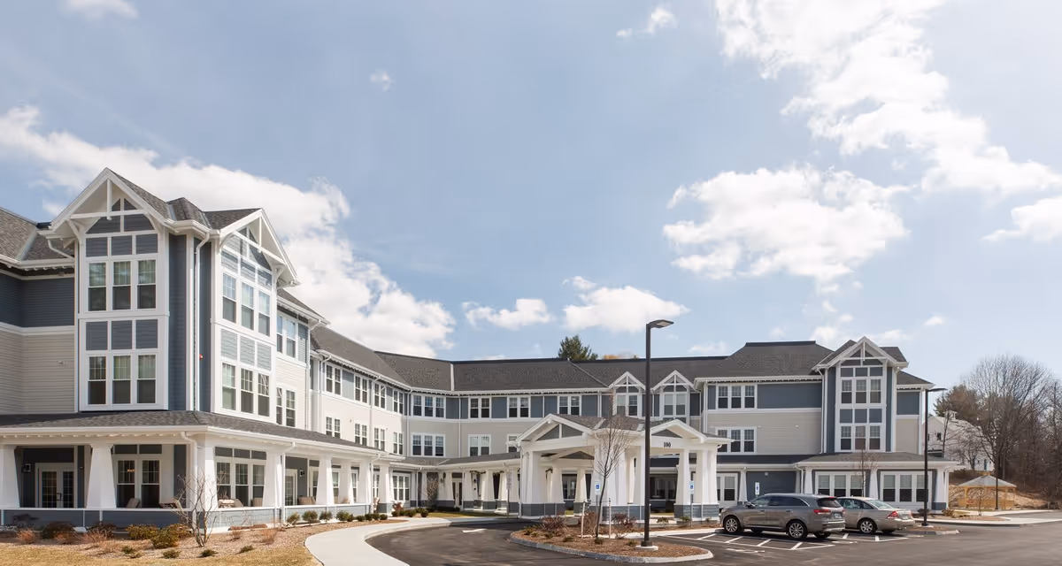 Front exterior of a multi-story senior living building with a covered entrance, parking lot, and cars under a partly cloudy sky.