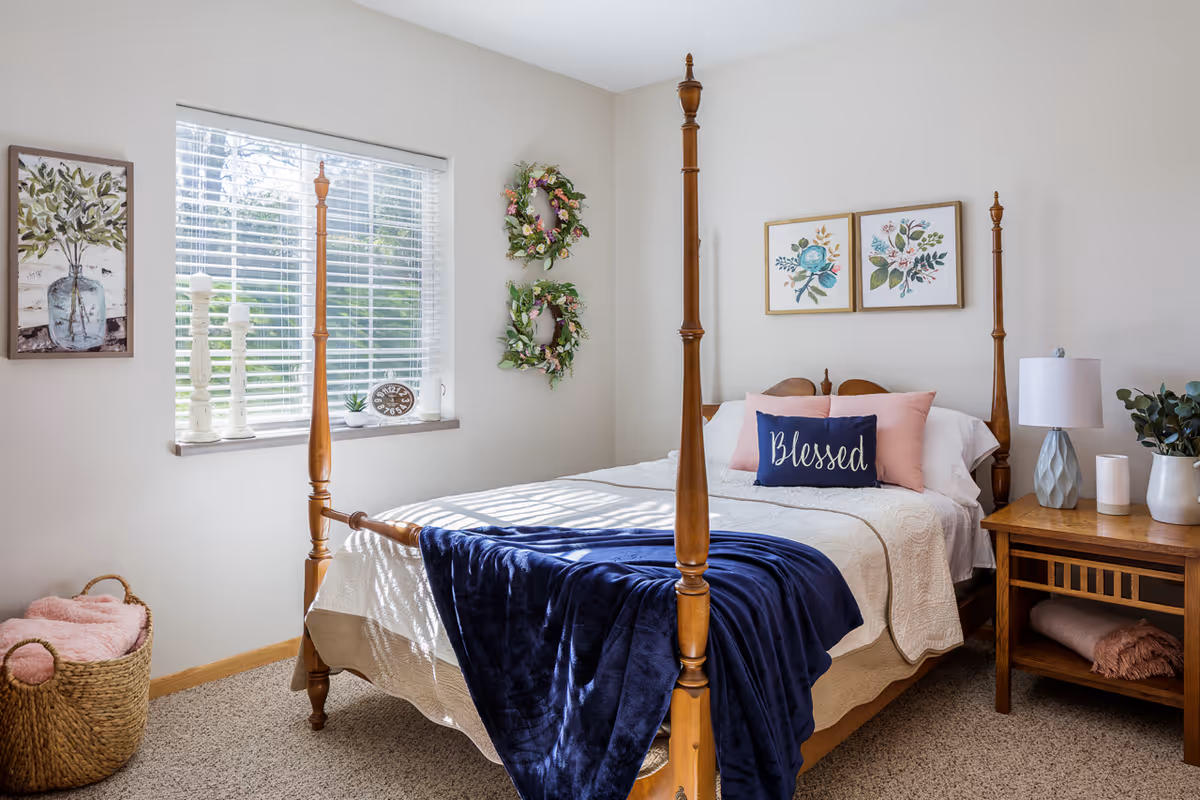 Bright bedroom featuring a wooden four-poster bed with white bedding, a navy throw and a 'Blessed' pillow, a window and wooden nightstand.