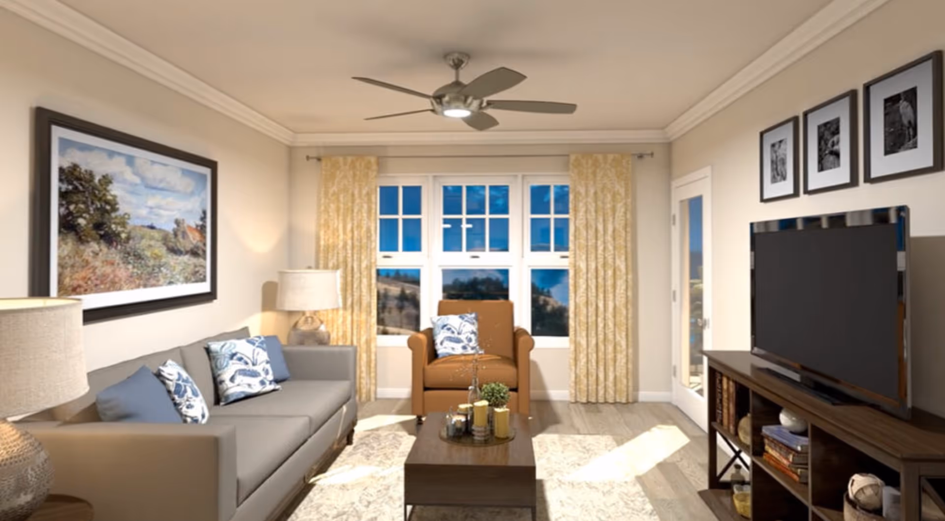 A cozy living room with a gray sofa adorned with blue and white patterned pillows, a brown armchair with a matching pillow, a wooden coffee table with decorative items, a large flat-screen TV on a wooden stand, three black and white framed pictures on the wall above the TV, a large landscape painting on the opposite wall, two table lamps, yellow patterned curtains framing a set of windows, and a ceiling fan with a light fixture.