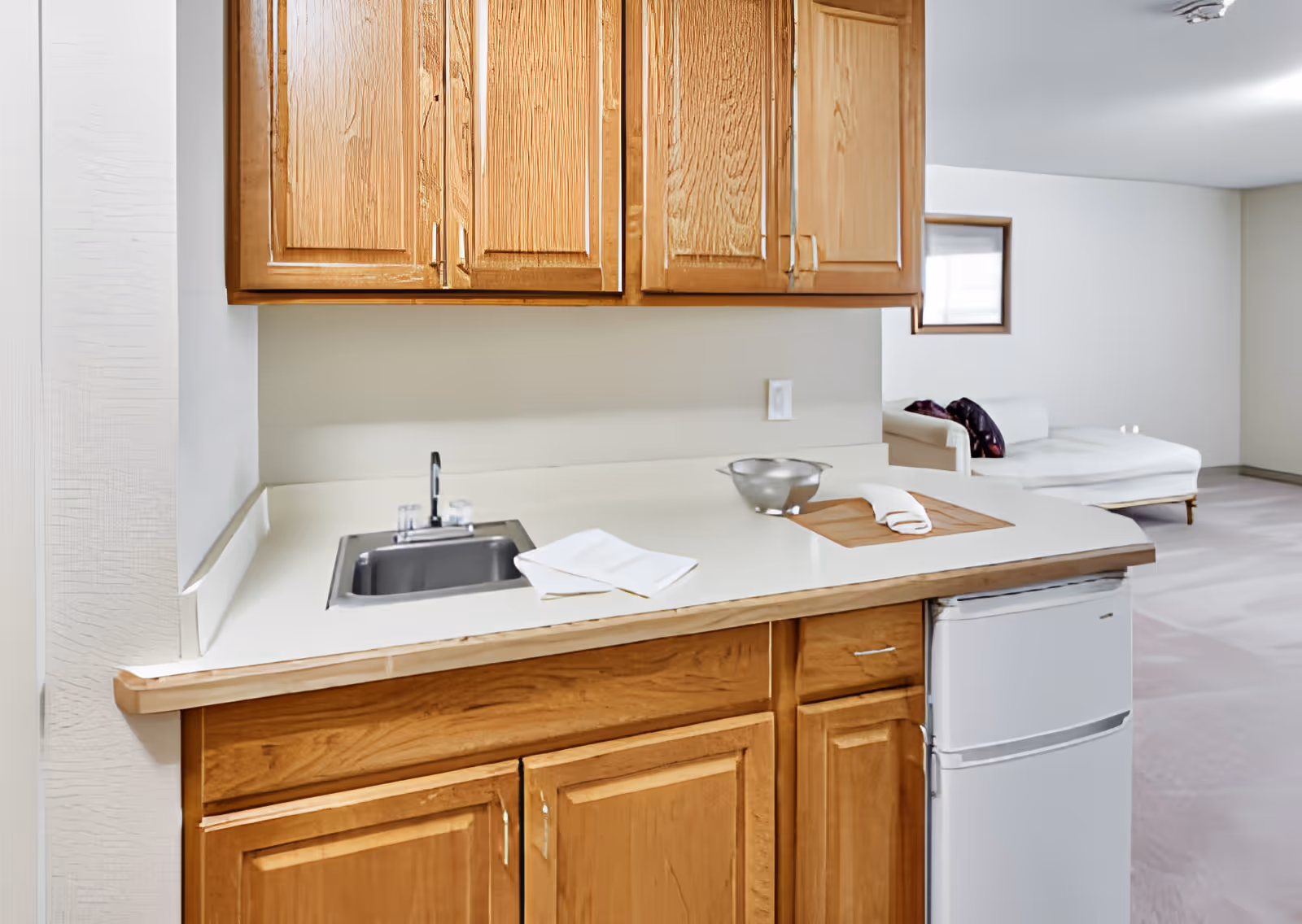 Small kitchenette with wooden cabinets, a sink and mini refrigerator, and a chaise lounge visible in the background.