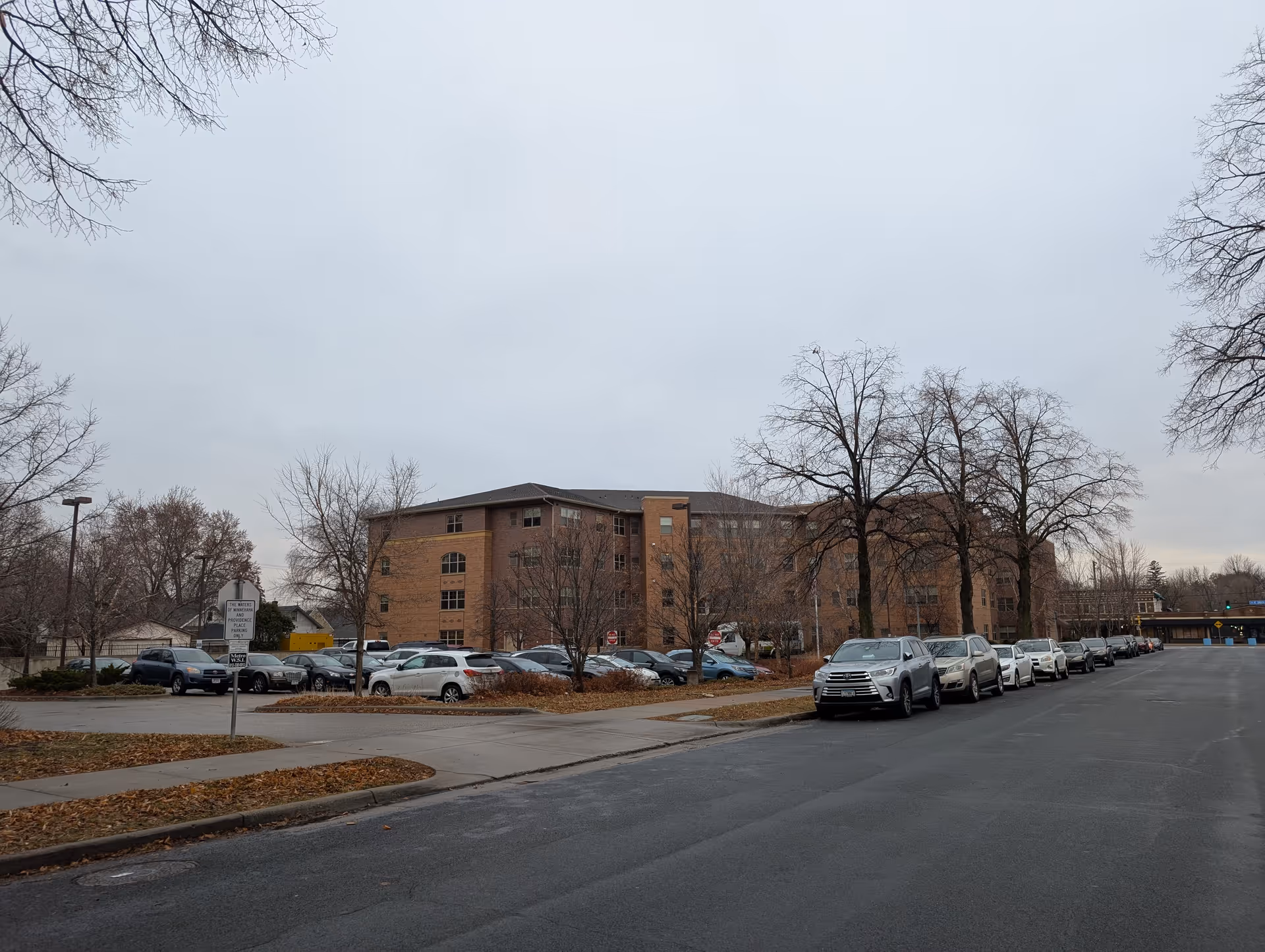 Exterior view of a multi-story brick building with a parking lot in front. Several cars are parked along the street and in the parking lot. Leafless trees and a cloudy sky are visible, indicating late fall or winter season.