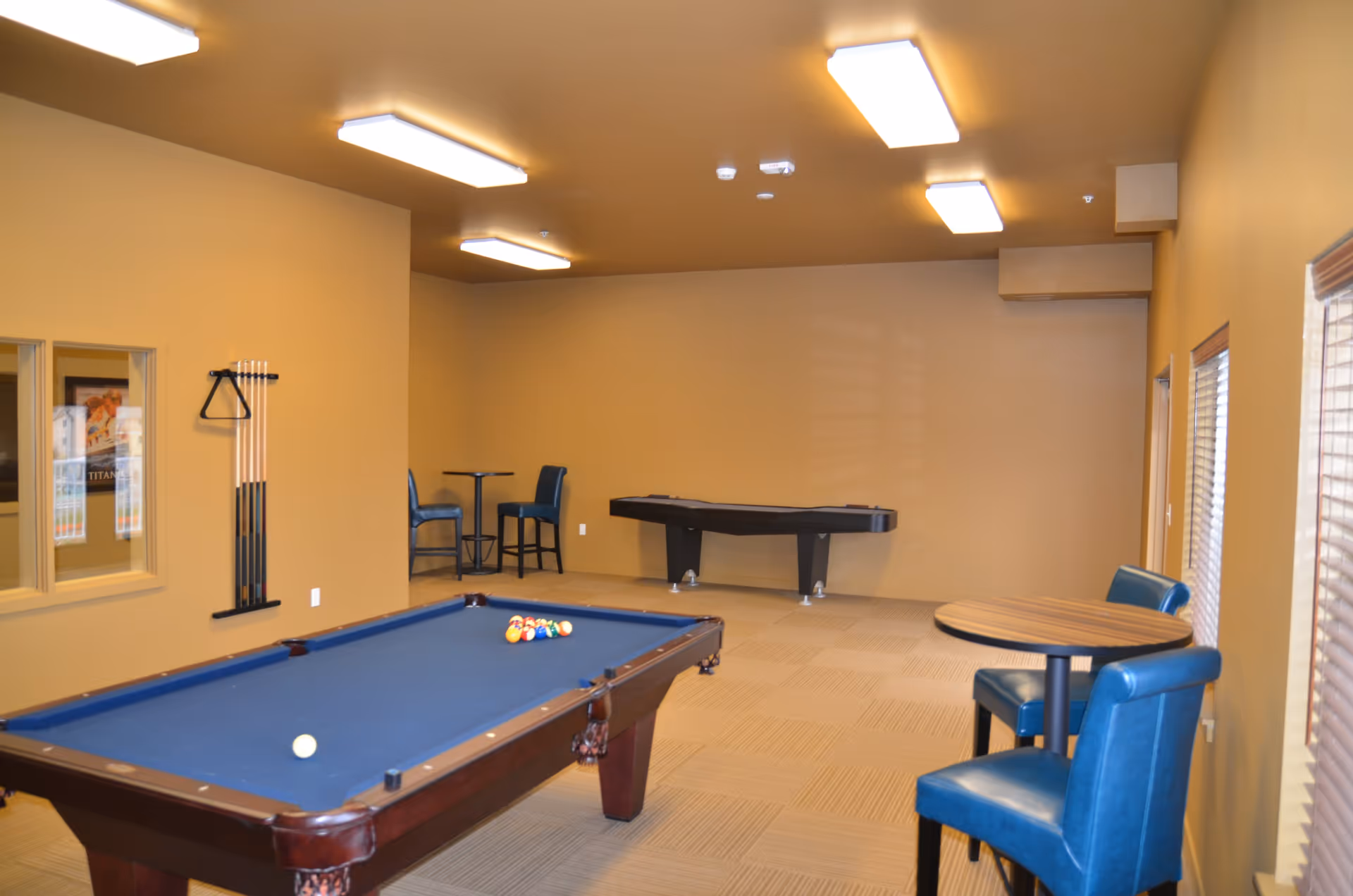 A game room with a blue felt pool table in the foreground, a wall-mounted rack holding pool cues, a small round table with two blue chairs near the window, and another small table with two blue chairs in the back corner. The room has beige walls, carpeted floor, and bright ceiling lights.