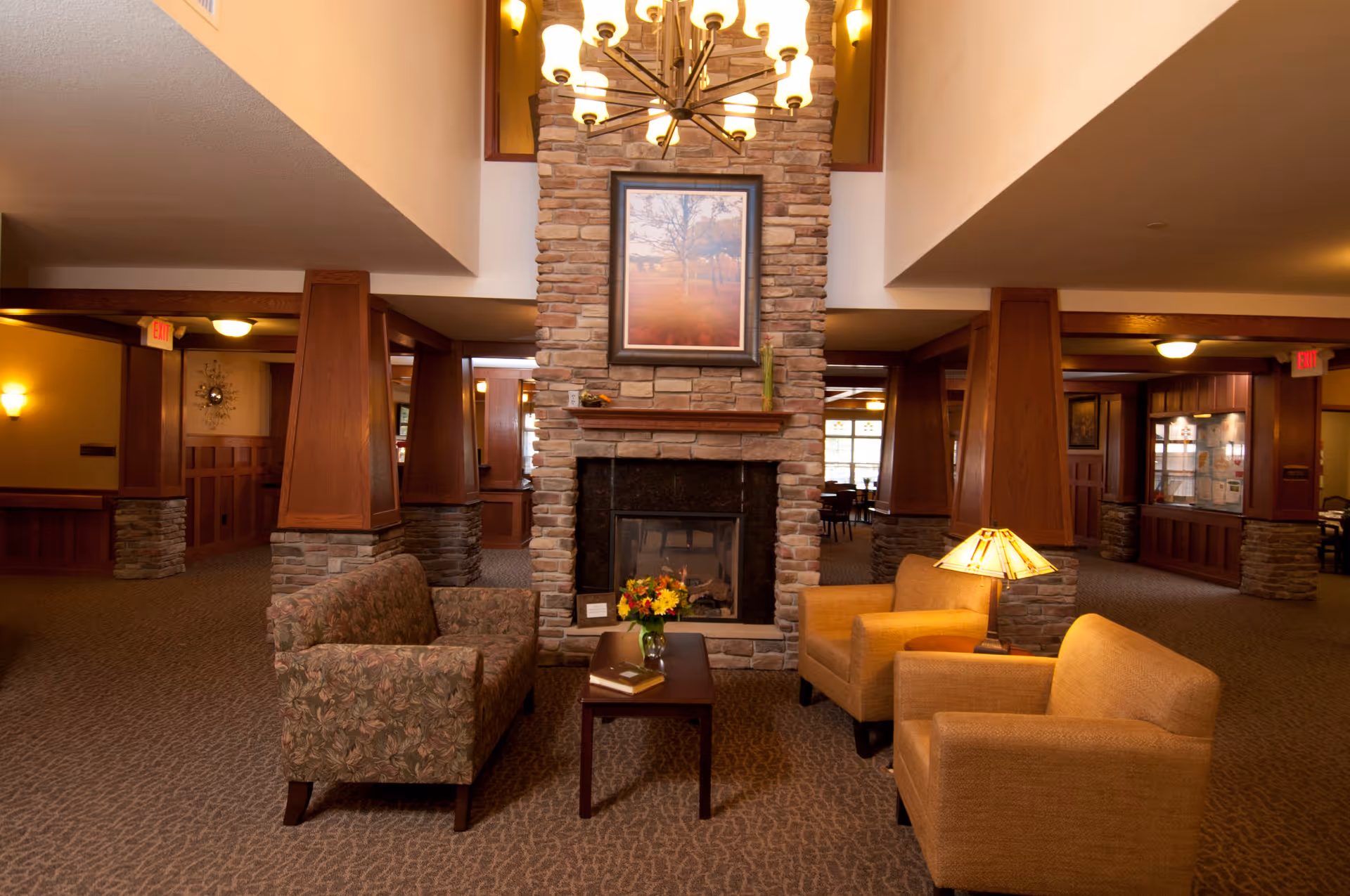 A cozy senior living facility common area with a stone fireplace in the center, flanked by a floral-patterned sofa on the left and two mustard-yellow armchairs on the right. A wooden coffee table with a vase of flowers and a book sits in front of the fireplace. Warm lighting and wooden pillars with stone bases enhance the inviting atmosphere.