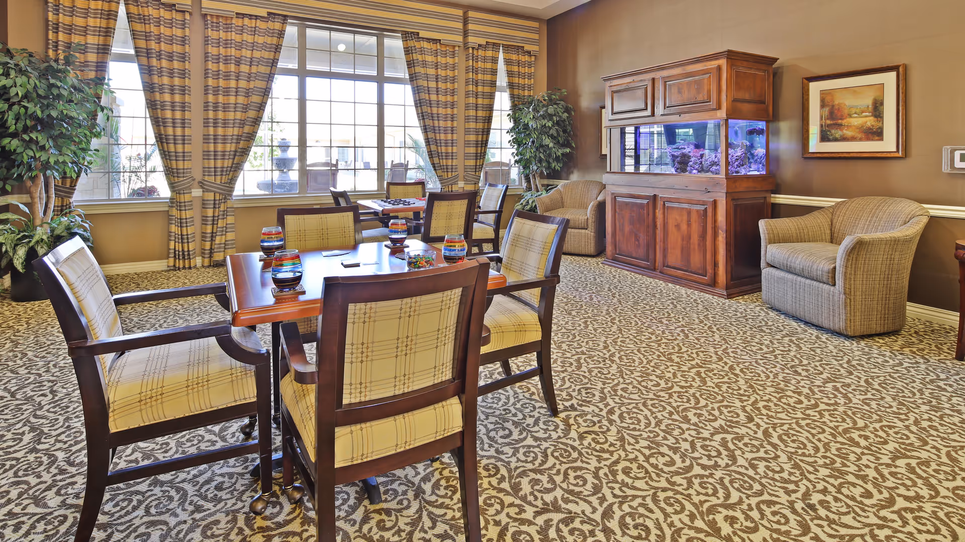 A well-lit common area with a patterned carpet, wooden table surrounded by six cushioned chairs with plaid upholstery, large windows with striped curtains, two armchairs, a large wooden cabinet with a built-in aquarium, potted plants, and a framed painting on the wall.