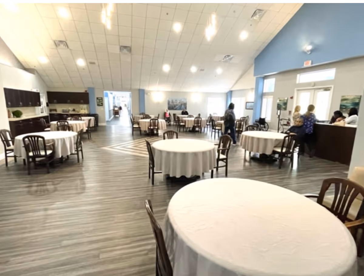Spacious dining/activity room with round tables covered in white tablecloths, wooden chairs, a high vaulted ceiling, and a reception area with a few people.