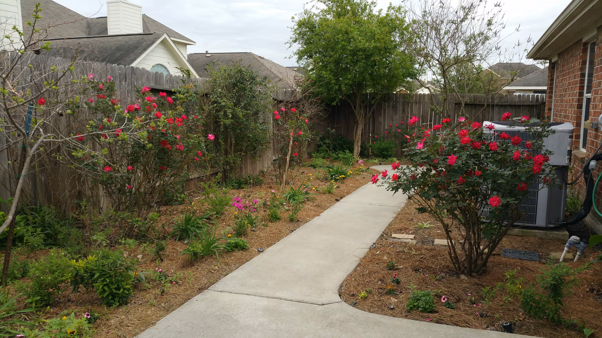 A garden pathway with blooming red and pink flowers along both sides, bordered by a wooden fence on the left and a brick building on the right. There is an air conditioning unit next to the building and some trees and shrubs in the background under a cloudy sky.