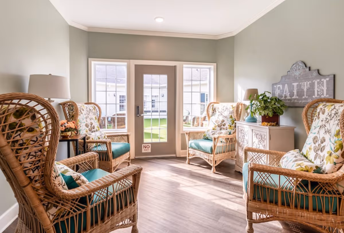 A cozy sitting area with four wicker armchairs featuring floral cushions arranged around a small white cabinet with a plant and lamp on top. The room has light green walls, wood flooring, and a door with windows on either side leading to an outdoor area with green grass. A decorative sign on the wall reads 'FAITH'.