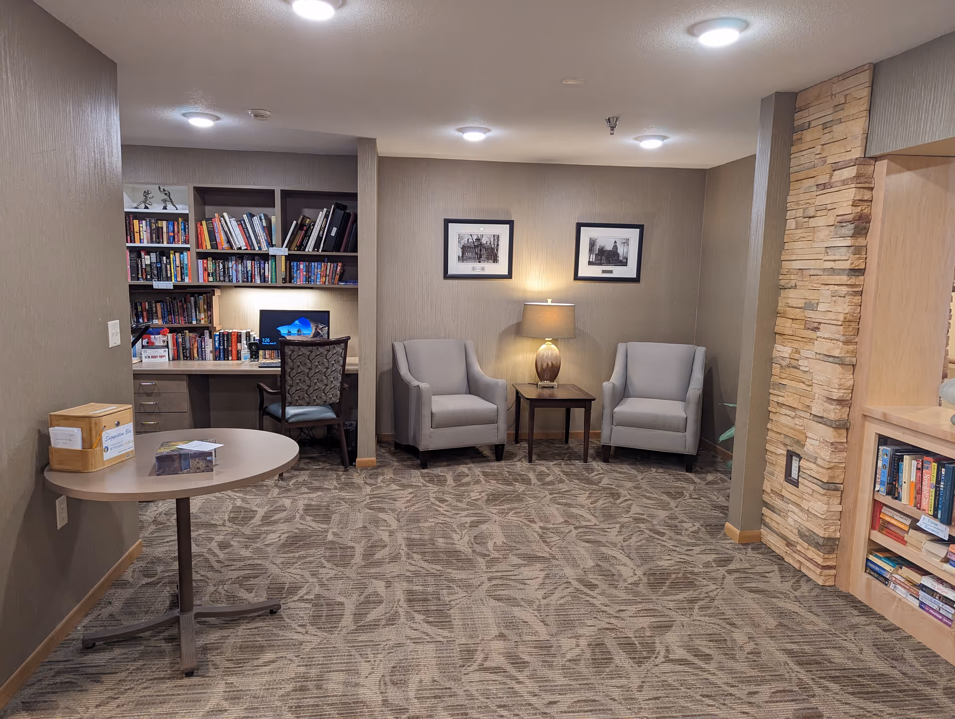 A cozy interior common area with two gray armchairs separated by a small wooden table with a lamp. Behind the chairs are two framed black and white pictures on the wall. To the left, there is a built-in desk with a computer and a chair, surrounded by bookshelves filled with books. A round table with a wooden box and some papers is in the foreground. The room has patterned carpet and warm lighting.