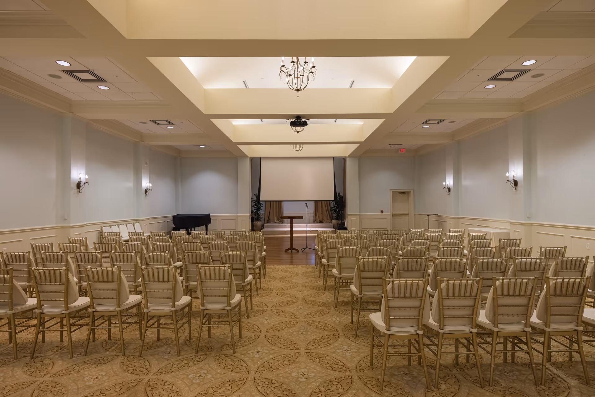 A large conference or event room with rows of beige chairs facing a stage area with a podium and a projection screen. The room has a patterned carpet, light blue walls with white wainscoting, wall sconces, and chandeliers hanging from a coffered ceiling. A black grand piano is visible to the left side of the stage.