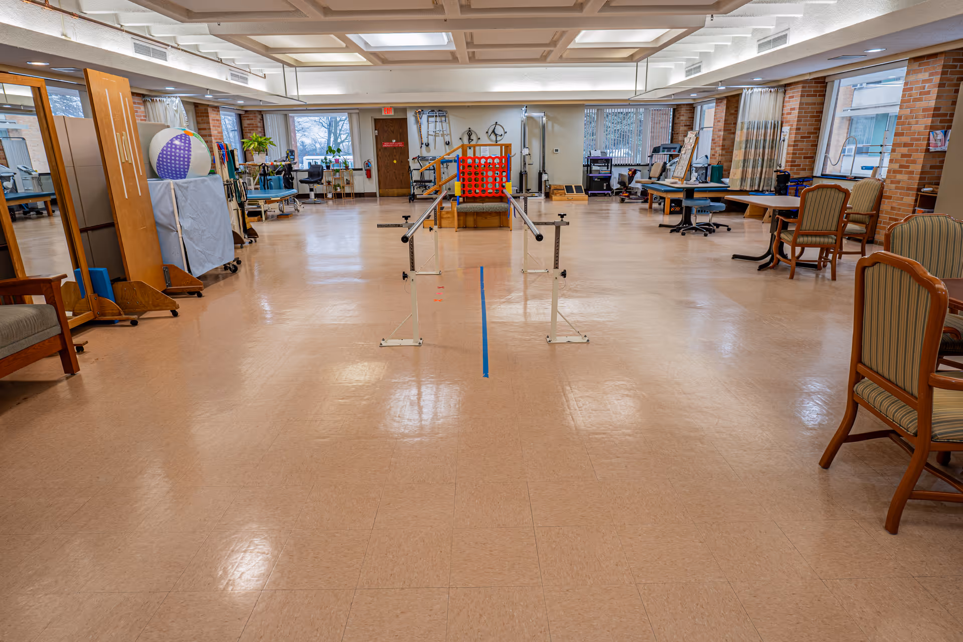 A spacious rehabilitation or physical therapy room with parallel bars in the center for walking exercises. The room has several chairs, tables, exercise equipment, and large windows letting in natural light. The floor is a light-colored tile, and the walls are brick with some curtains on the windows.