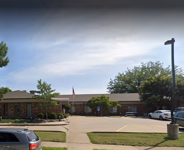 Exterior view of a single-story brick senior living facility with parking and trees.