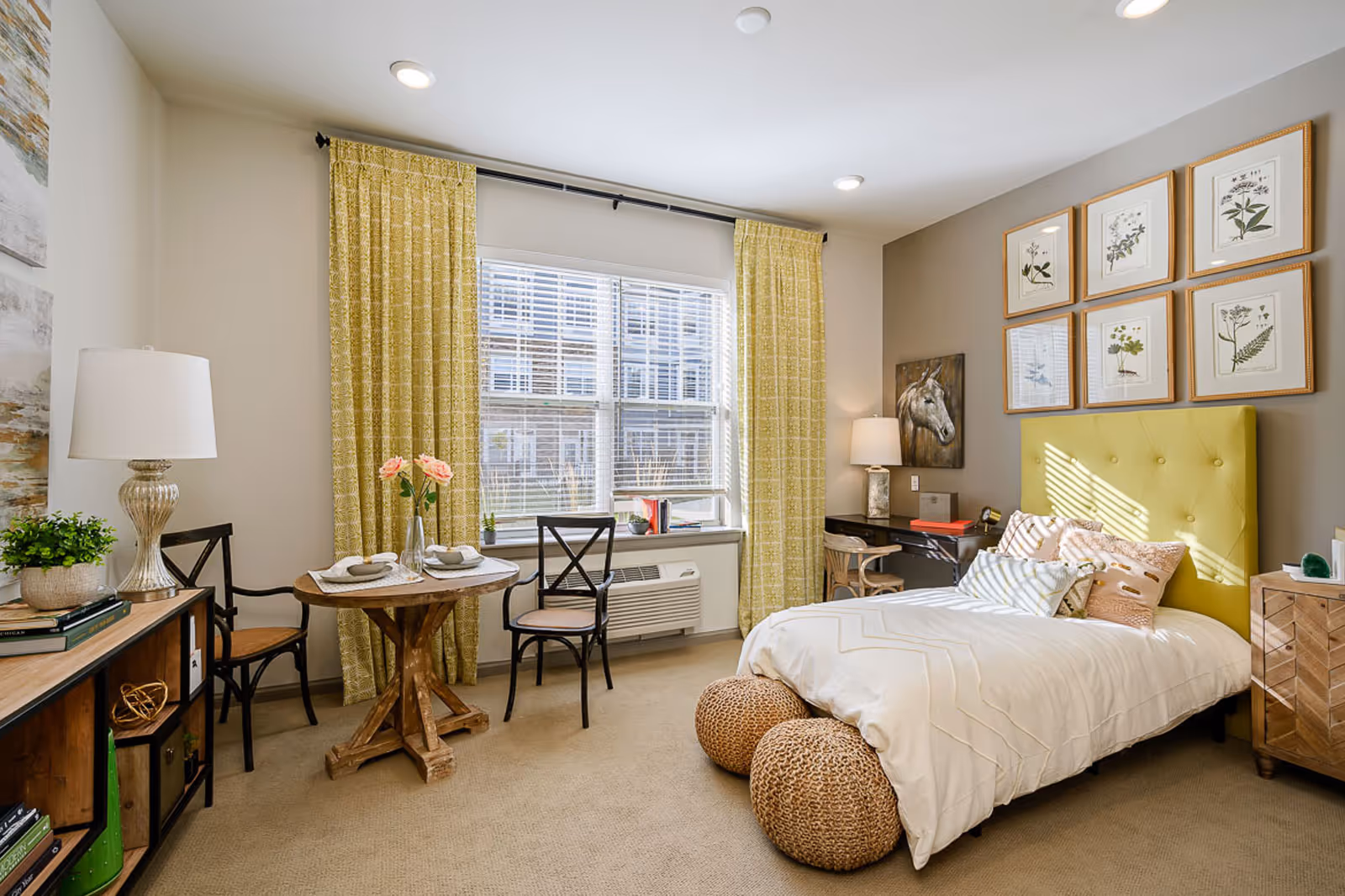 A bright and cozy bedroom with a large window covered by yellow patterned curtains. The room features a bed with a yellow tufted headboard, white bedding, and several decorative pillows. Two woven poufs are placed at the foot of the bed. There is a small wooden table with two chairs near the window, set with plates and napkins. A wooden dresser with a lamp and plants is on the left side, and a desk with a lamp and chair is on the right side. The walls are decorated with framed botanical prints and a horse painting.