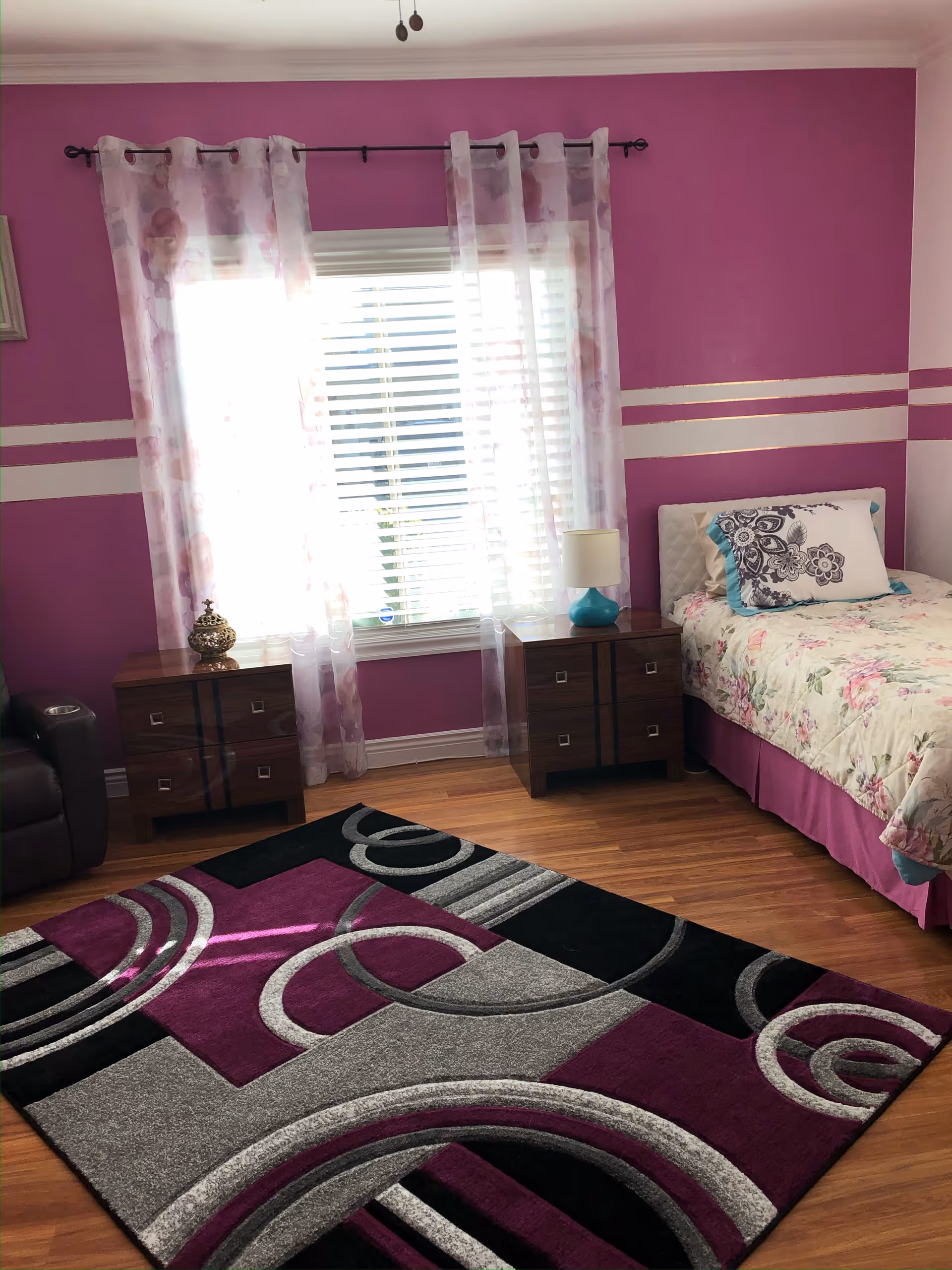 A bedroom with bright purple walls featuring two white horizontal stripes. There is a window with white blinds and sheer floral curtains. On either side of the window are two dark wooden nightstands, one with a decorative item and the other with a lamp with a turquoise base. A single bed with floral bedding and a decorative pillow is positioned to the right. The floor is wooden with a large area rug in shades of purple, black, and gray with circular patterns.