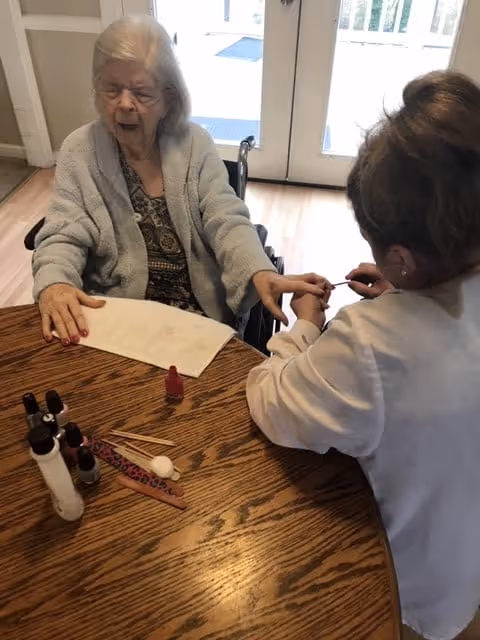 An elderly woman in a wheelchair is having her nails painted by a caregiver at a wooden table. Various nail care items, including nail polish bottles and a nail file, are on the table. The setting appears to be indoors with glass doors in the background.