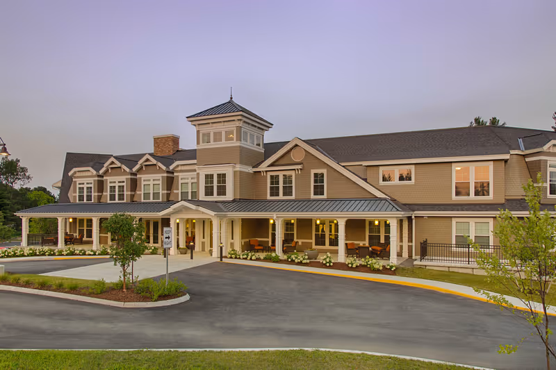 Exterior view of a large two-story senior living facility building with a covered entrance, multiple windows, and a tower-like structure in the center. The building is surrounded by landscaped greenery and a paved driveway.