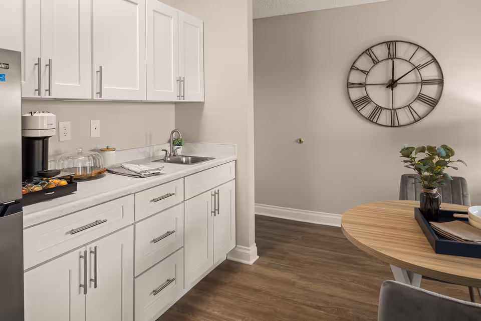 A modern kitchen area with white cabinets and drawers, a countertop with a sink, a coffee maker with coffee pods, and a glass cake dome. Adjacent to the kitchen is a round wooden dining table with a black tray holding a bowl and napkins, a small plant in a vase, and gray upholstered chairs. A large decorative wall clock with Roman numerals hangs on the beige wall above the dining area. The floor is wood with a medium brown finish.