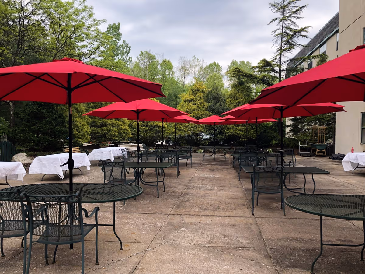 Outdoor patio area with multiple black metal tables and chairs, each shaded by large red umbrellas. The patio is surrounded by green trees and bushes, with a building visible on the right side. Several tables covered with white tablecloths are placed along the edges of the patio.