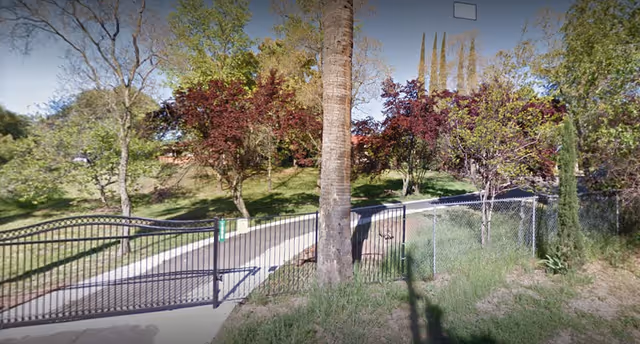 A gated entrance with a black metal fence and gate, surrounded by trees with green and reddish leaves, and a paved pathway leading uphill.