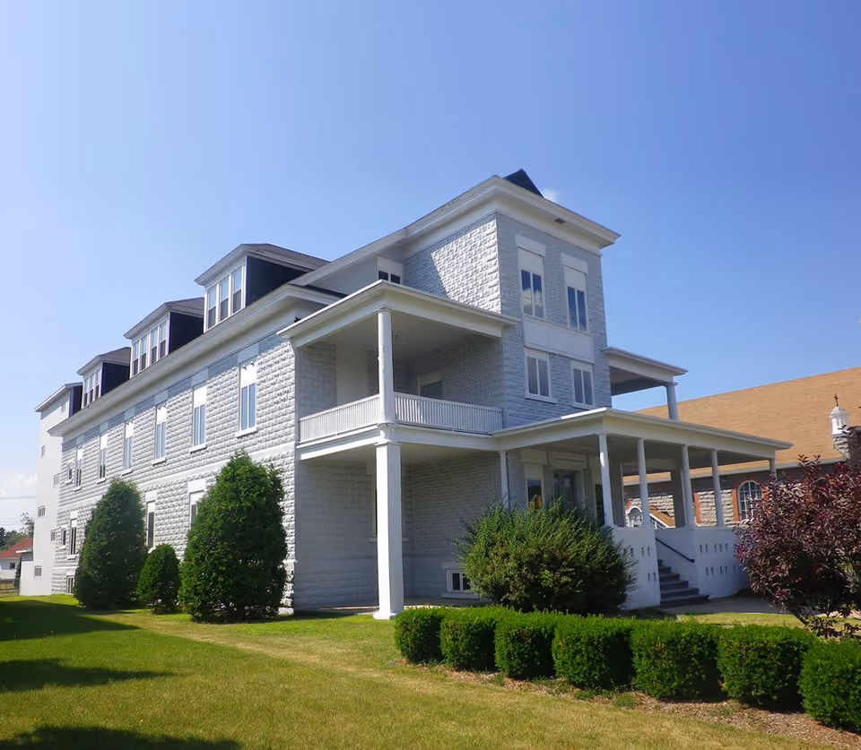 A large, multi-story white building with a wrap-around porch and multiple windows, surrounded by green bushes and trees under a clear blue sky.