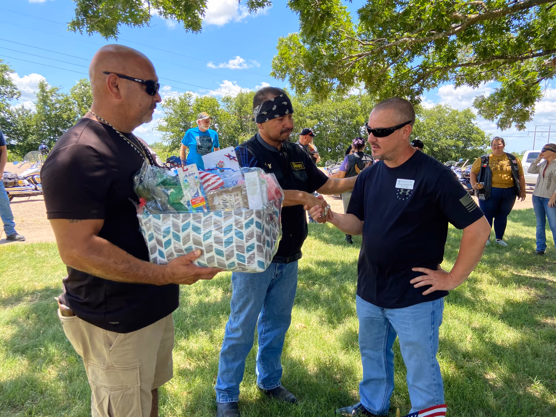 Three men outdoors on a sunny day, two of them shaking hands while the third holds a gift basket filled with various items. Other people and motorcycles are visible in the background under a tree with green leaves.