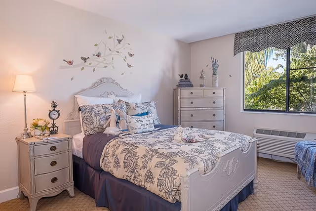 A cozy bedroom with a vintage-style white bed frame and floral bedding. The bed is neatly made with multiple pillows and a stuffed animal. To the left of the bed is a small nightstand with a lamp, a clock, and a flower arrangement. On the wall above the bed is a decorative decal of birds on branches. To the right is a tall dresser with decorative items on top. A window with a patterned valance shows green trees outside. The room has beige walls and carpeted floor.