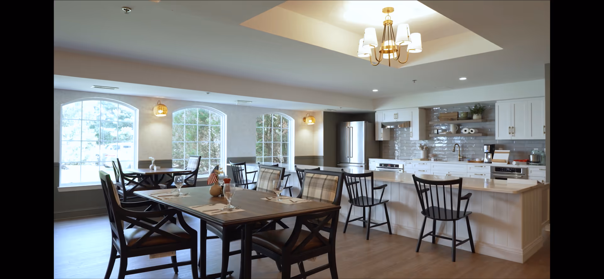 A bright and spacious dining area with large arched windows letting in natural light. The room features a wooden dining table set with glassware and napkins, surrounded by chairs with plaid cushions. In the background, there is a modern kitchen with white cabinetry, a stainless steel refrigerator, a coffee maker, and a kitchen island with bar stools. The ceiling has a recessed light fixture with a chandelier.
