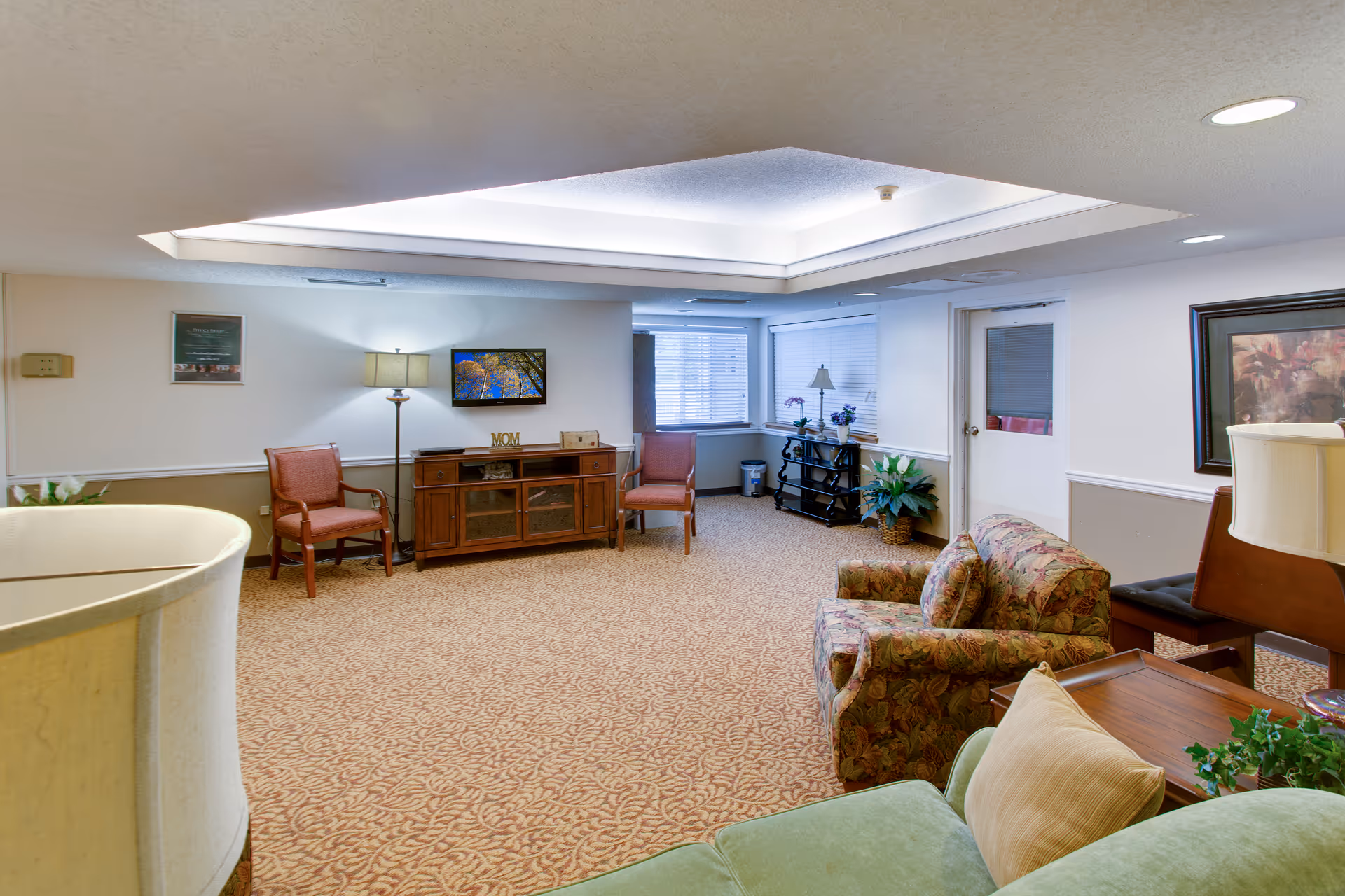 A well-lit senior living common room with upholstered chairs and sofas, a TV on a wooden cabinet, lamps, and patterned carpet.