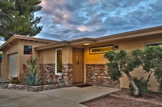 Front entrance of a single-story care home with a brick-and-stucco facade, covered doorway, driveway, and landscaping under a cloudy sky.