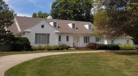 A large white single-story house with a sloped roof and multiple dormer windows, surrounded by green grass, trees, and shrubs under a blue sky.