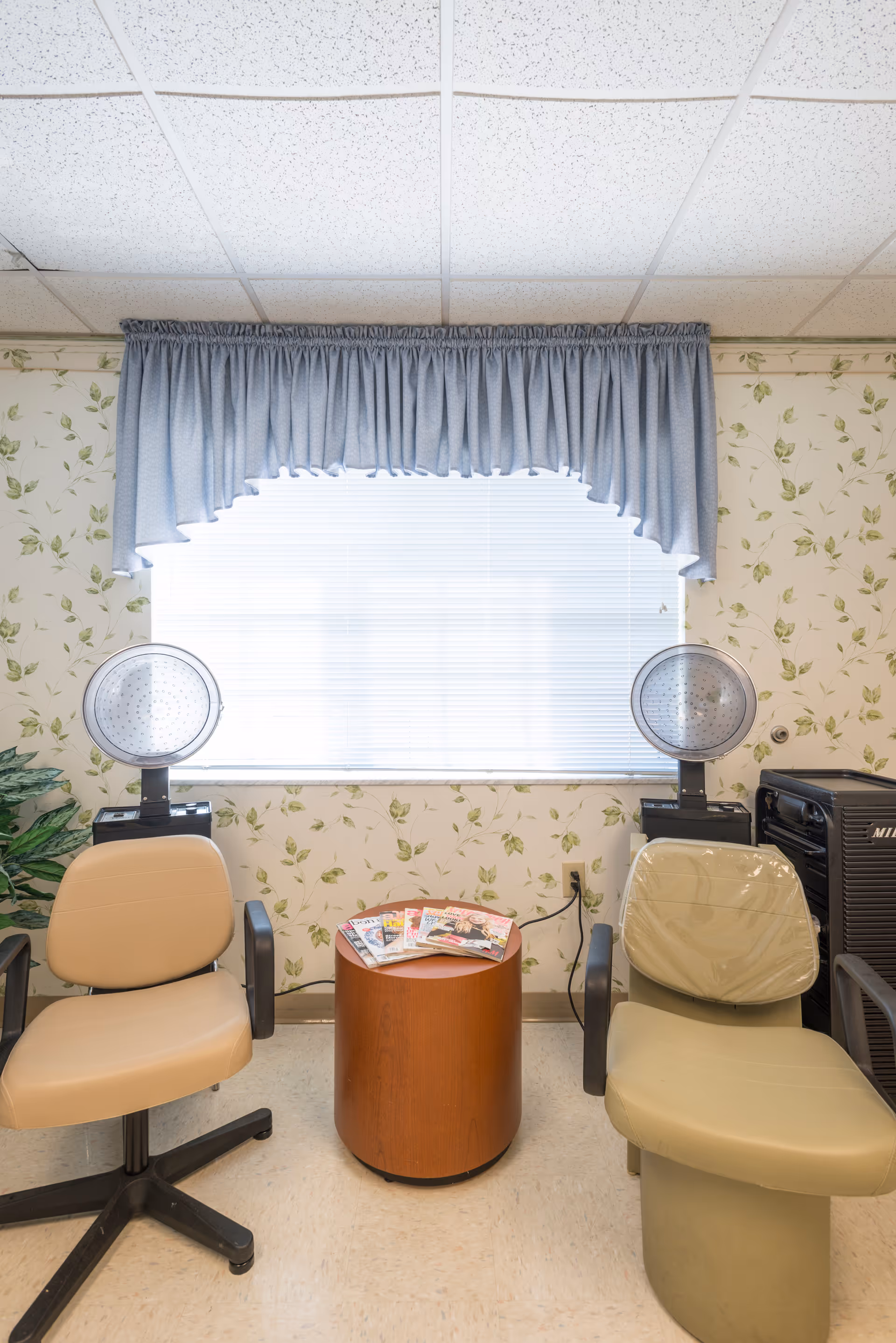 A small room with two beige salon chairs facing forward, each with a hair dryer hood behind them. Between the chairs is a small round wooden table with magazines on top. The wall behind has floral wallpaper and a window with closed blinds and a blue valance curtain.