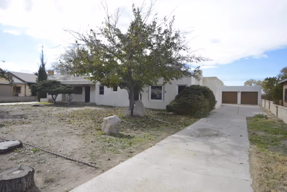 Single-story white stucco building with a tree in the front yard and a concrete driveway leading to a two-car garage.