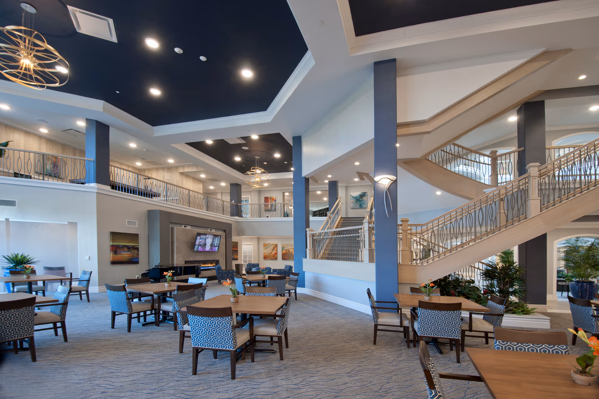 Spacious and well-lit senior living facility common area with multiple tables and chairs arranged for dining or socializing. The room features a high ceiling with modern light fixtures, a staircase with decorative railings, and a fireplace with a mounted television above it. There are plants and artwork decorating the space, creating a welcoming atmosphere.