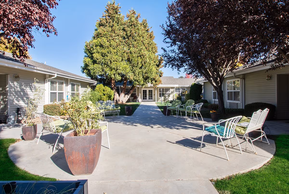 Outdoor courtyard area at Avista Senior Living Nampa with metal chairs and benches arranged on a concrete patio surrounded by green grass, potted plants, and trees. Single-story buildings with windows and doors border the courtyard under a clear blue sky.