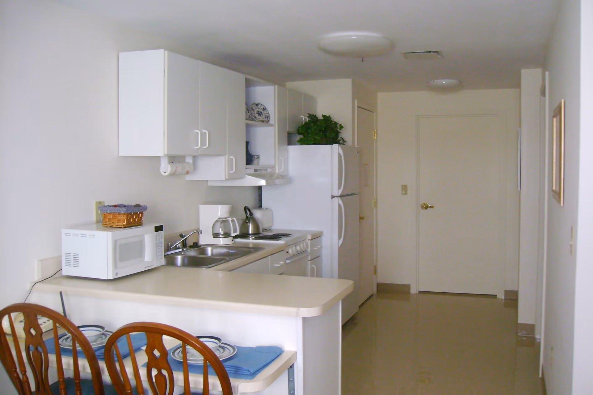 A small kitchen area with white cabinets, a white refrigerator, a microwave, a coffee maker, a kettle, and a toaster. There is a counter with two wooden chairs set with plates and blue napkins. The floor is shiny and beige, and the walls are white.